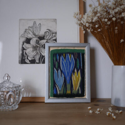 Framed colorful woven textile on a wooden shelf beside dried flowers and a glass jar, with a floral sketch on the wall behind.