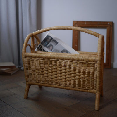 Wicker basket on a wooden floor with a folded newspaper inside and a wooden frame leaning against the wall in the background.