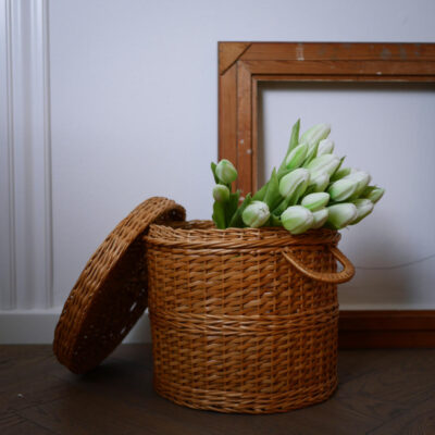 Wicker basket with its lid leaned against the side, filled with a bouquet of white tulips on a dark wooden floor.