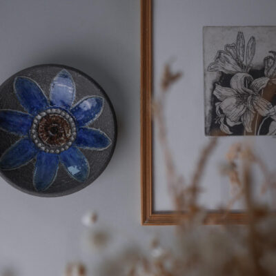 Blue ceramic vase with a six-petal flower motif next to a framed botanical print on a wall, decorative still life arrangement