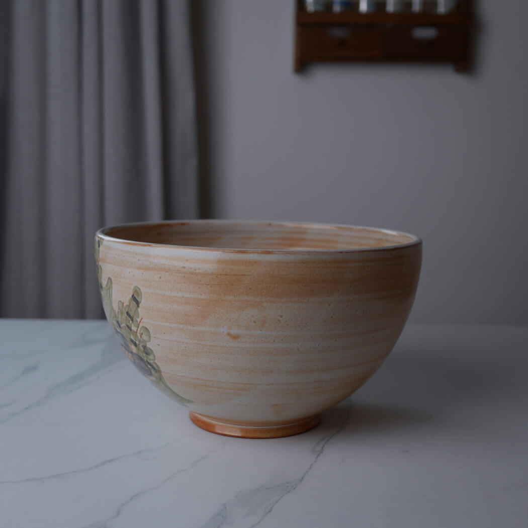 Ceramic bowl with orange-tan glaze and green leaf pattern sits on a white marble table in a softly lit room.