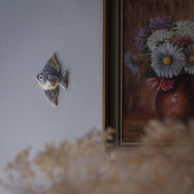 Ceramic fish wall ornament on a light gray wall beside a framed floral painting showing daisies in a vase, with dried flowers in the foreground.