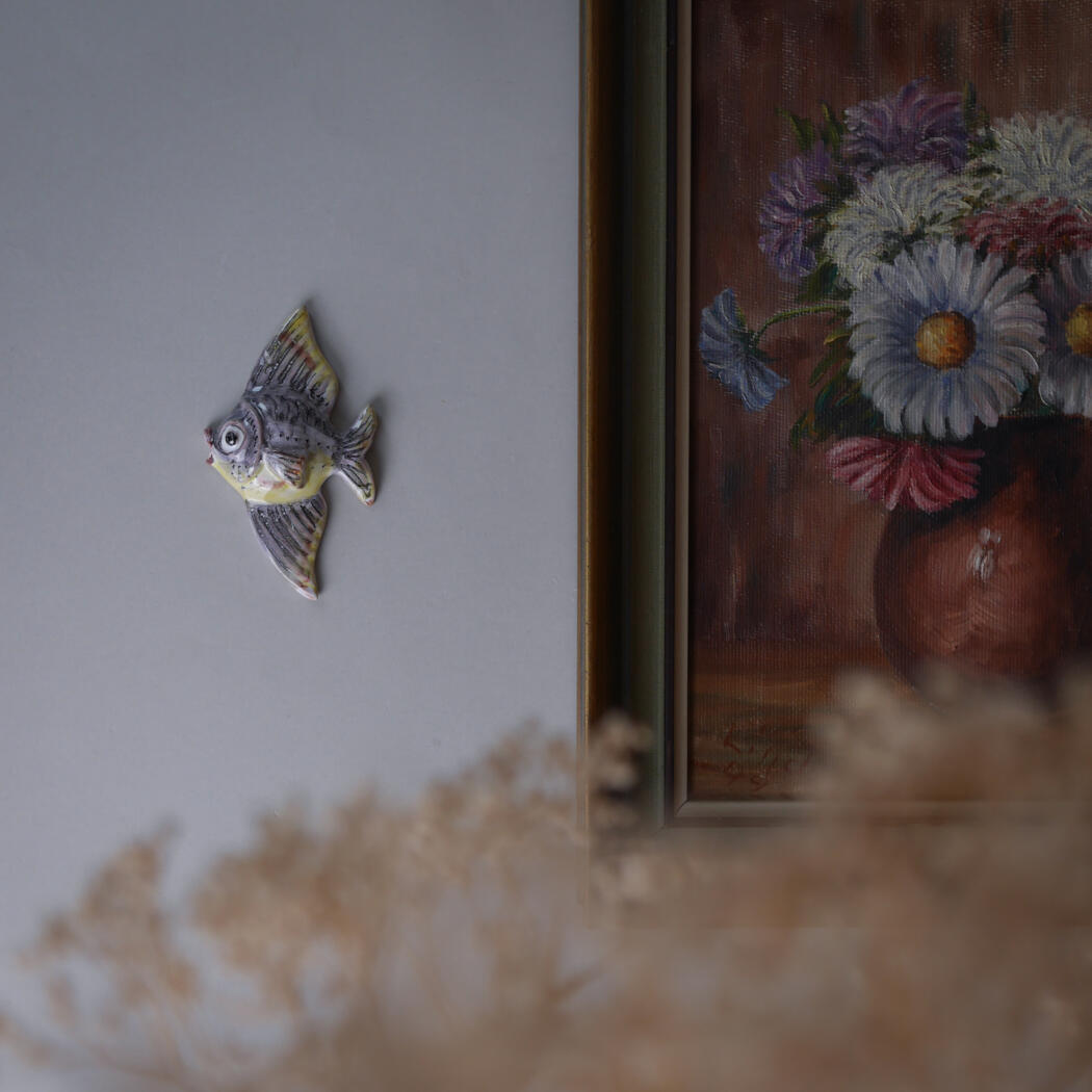 Ceramic fish wall ornament on a light gray wall beside a framed floral painting showing daisies in a vase, with dried flowers in the foreground.
