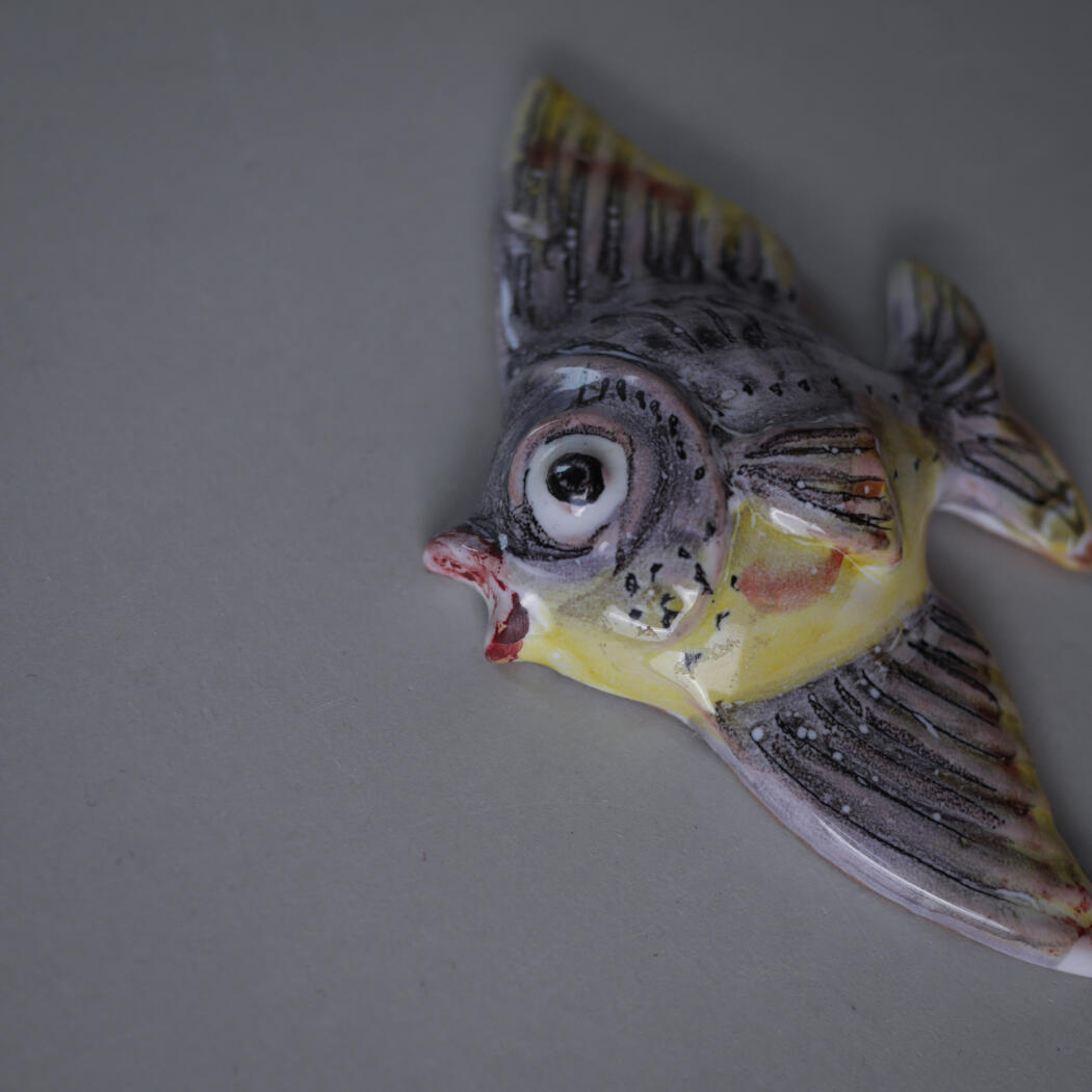 Colorful ceramic fish head with a large eye, open mouth, and yellow-gray pattern on a plain gray surface.