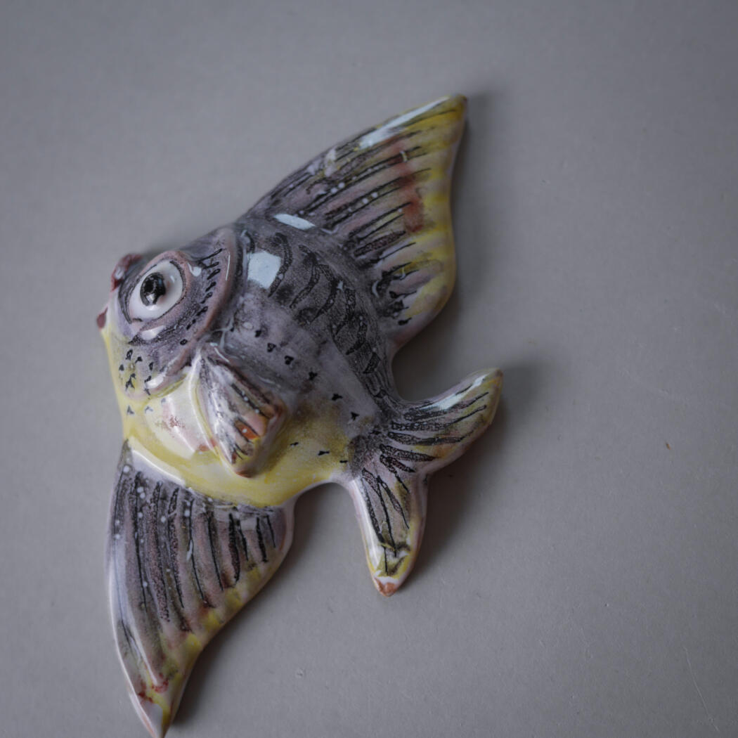 Ceramic flying-fish ornament with gray body and yellow accents, detailed black fins, lying on a plain surface