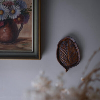 Framed painting of colorful flowers on a grey wall beside a brown leaf-shaped ceramic wall ornament.