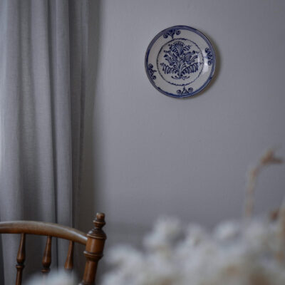 Blue and white decorative plate on a light gray wall, with gray curtains to the left and a wooden chair in the foreground.