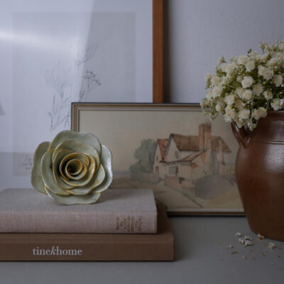 Decorative still life: a ceramic pale-green rose rests on neutral books beside a vase of white flowers and a framed countryside painting in the background.