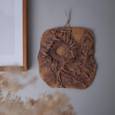 Decorative clay plaque with a sunflower relief hanging on a gray wall by a twine cord near a wooden frame.
