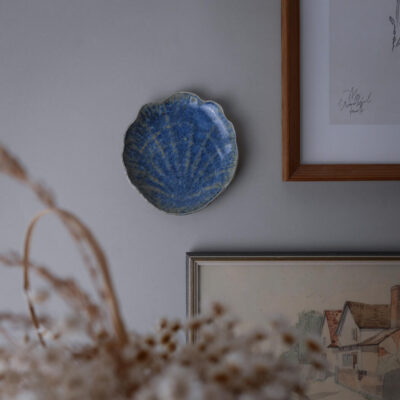 Blue scalloped ceramic dish mounted on a gray wall, with framed artwork nearby and dried flowers in the foreground.