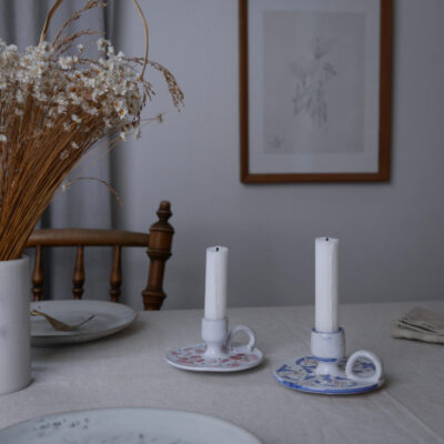 A dining table with two decorative ceramic candle holders and white candles, dried flowers in a vase on the left, and a framed sketch on the wall behind.