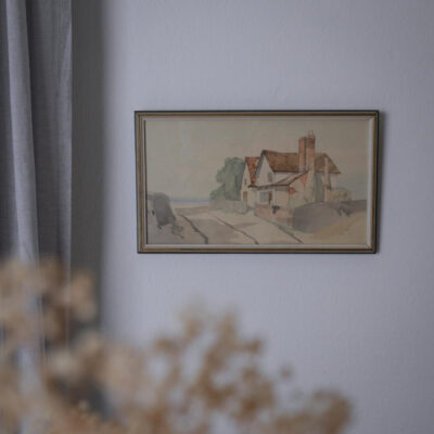 Framed watercolor painting of a cottage with a red roof on a country road, hung on a light wall with a curtain to the left.