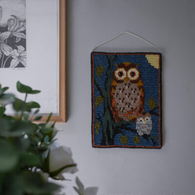 Woven wall hanging showing a brown owl and a small white owl on a blue background, hanging on a gray wall near a frame.