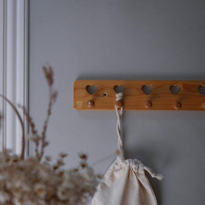 Wooden wall-mounted coat rack with heart-shaped cutouts and a drawstring bag hanging from a rope above a grey wall.