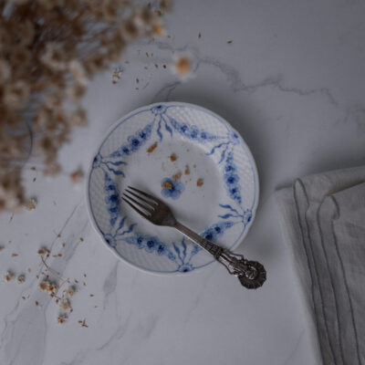Blue-and-white patterned plate with a few crumbs and a fork on a marble countertop, dried flowers to the left, and a folded gray napkin to the right.