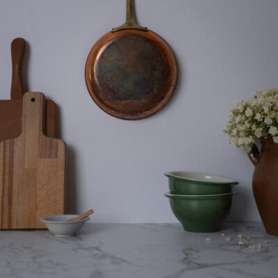 Copper frying pan hanging on a light wall above a marble countertop with wooden cutting boards to the left and a vase of white flowers on the right.