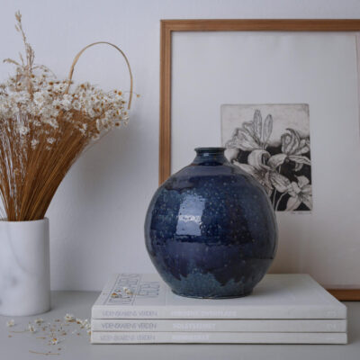 Blue round ceramic vase on stacked white books with dried flowers in a white vase and a framed botanical print behind.