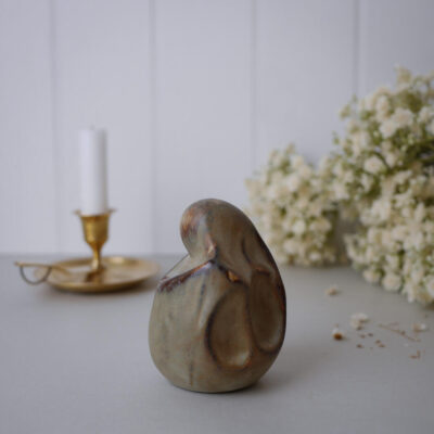 Abstract ceramic sculpture with a rounded, organic form on a pale tabletop, blurred candle holder and white flowers in the background.