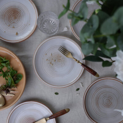 Top-down view of a table setting with white plates that have orange rims, a fork resting on one plate, and a glass nearby on a light linen tablecloth with a beige salad plate and greens visible at the edge.