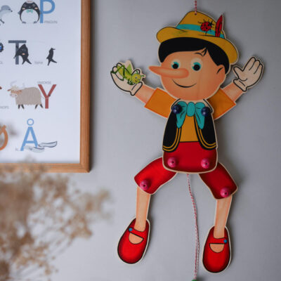 Hanging cartoon Pinocchio puppet with yellow hat and red shoes beside a framed alphabet poster on the wall.