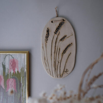 Oval ceramic wall plaque with pressed wheat stalks hanging by twine on a gray wall
