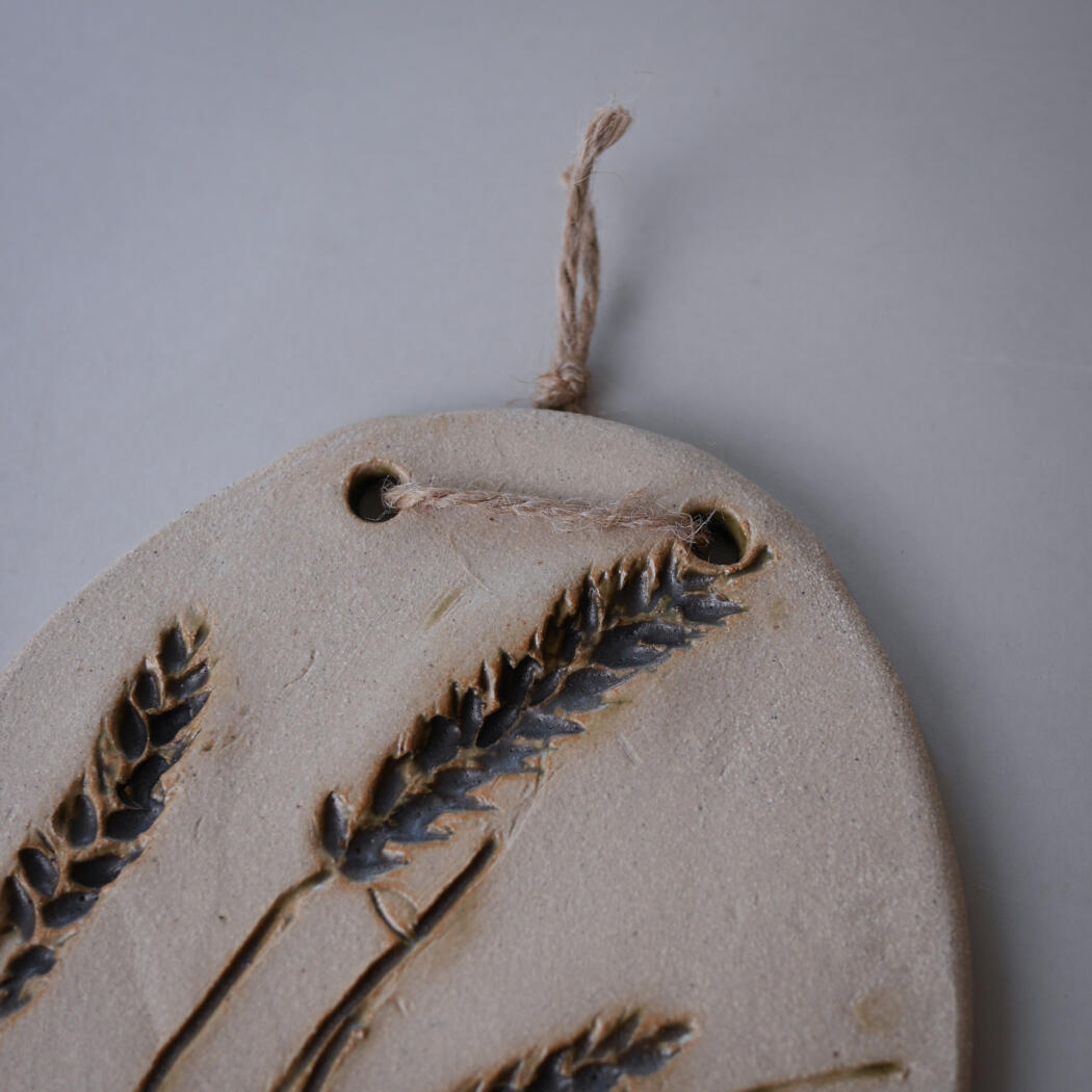 Round ceramic ornament with pressed lavender/wheat-like stalks and a hanging jute loop for wall display.