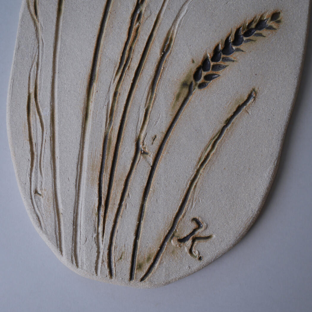 Decorative ceramic plaque with etched grass stalks and a wheat head on a light, oval surface.