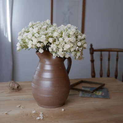 Brown ceramic pitcher filled with small white flowers on a wooden table, with a few petals scattered nearby.