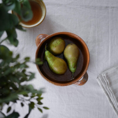 Top-down view of a terracotta pot with three poached pears in dark wine sauce on a light table, with a cup and blurred green plant nearby.
