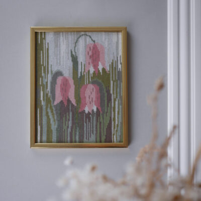 Framed cross-stitch art of pink flowers hanging on a gray wall with a gold frame.