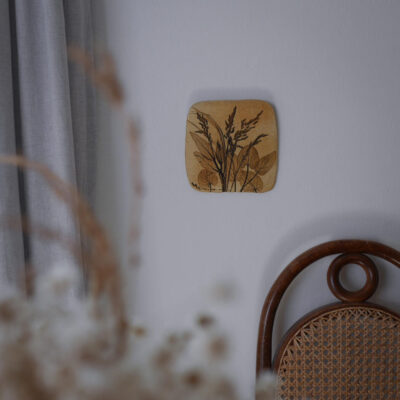 Small square wall art depicting dried grasses, mounted on a light wall above a wicker cane chair.
