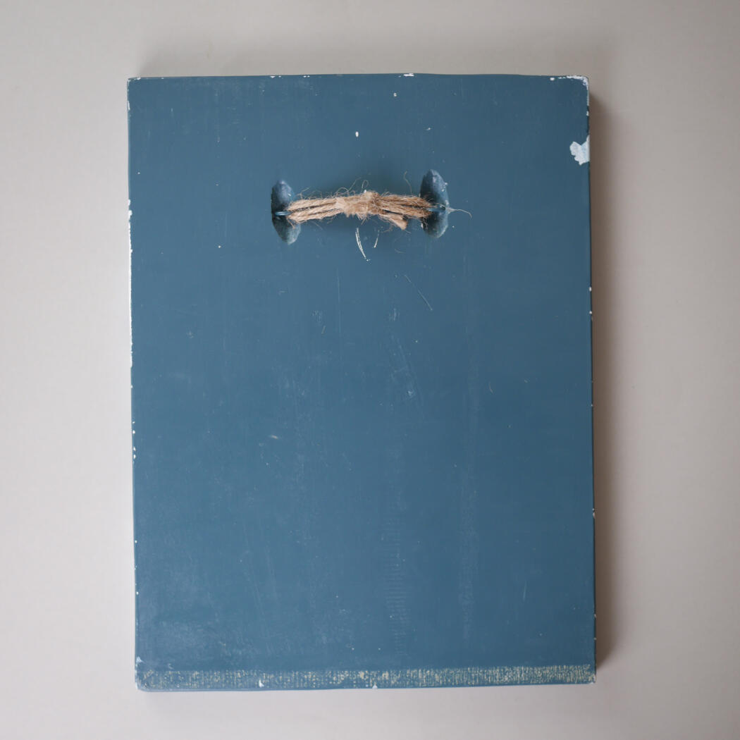 Blue wooden panel with a frayed natural rope threaded through two dark eyelets at the top center, worn and weathered.
