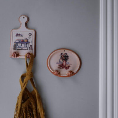 Two ceramic wall hooks: a pink kitchen-themed plaque with a mustard bag hanging from it, beside a round peach-colored plate featuring a floral vase design on a gray wall.