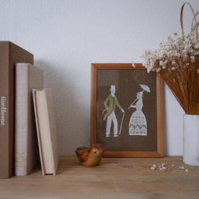 Framed textile embroidery of a man with a cane and a woman in a dress, on a brown background, on a wooden shelf beside dried flowers and a wooden bird.