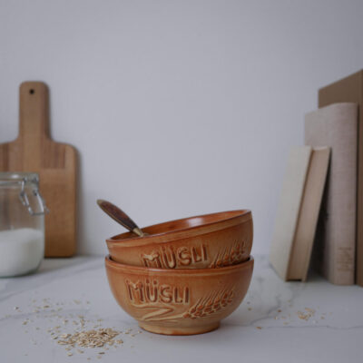 Two brown ceramic bowls embossed with 'Müsli' stacked on a white countertop, a spoon resting inside, oats scattered nearby in a kitchen scene.