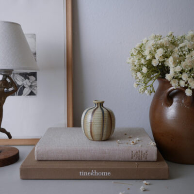 Still-life scene with a striped ceramic vase on a beige book atop a larger brown book that reads 'tinekhome', flanked by a brown vase of white flowers on the right and a lamp on the left.