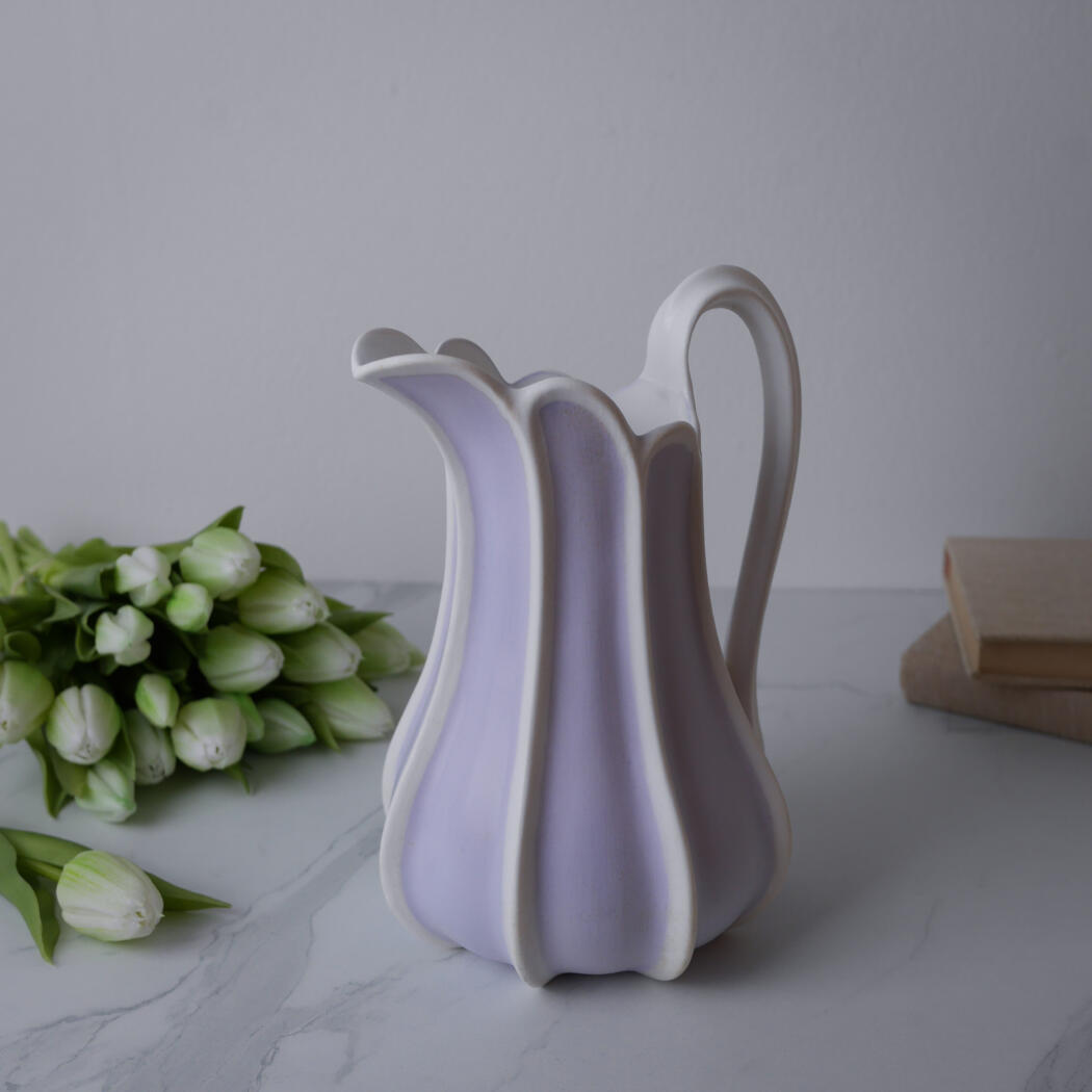 Lavender ceramic vase with white vertical ridges on a marble surface, with white tulips nearby and a stack of books in the background.
