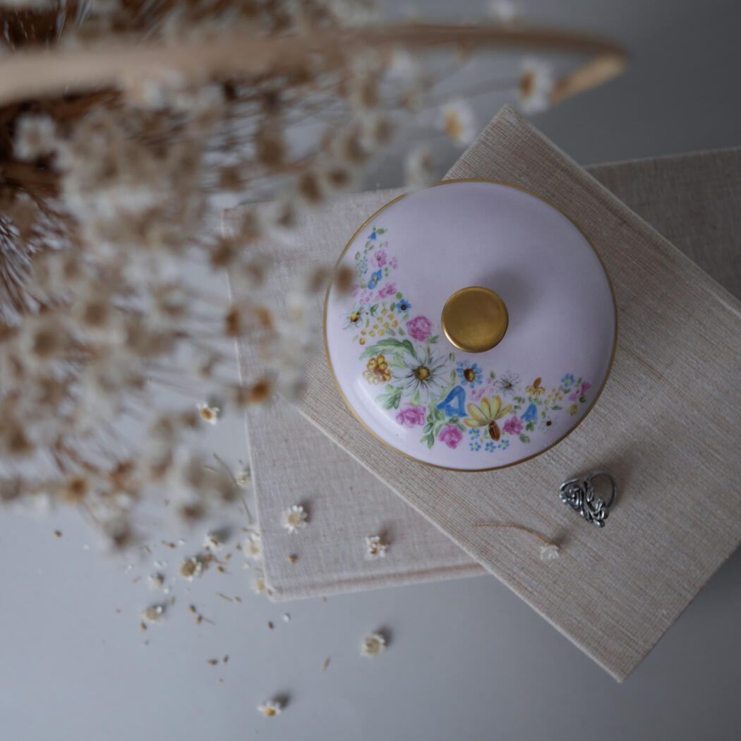 Round porcelain lid with gold knob and floral pattern resting on beige napkins, surrounded by dried flowers and a small silver heart charm.