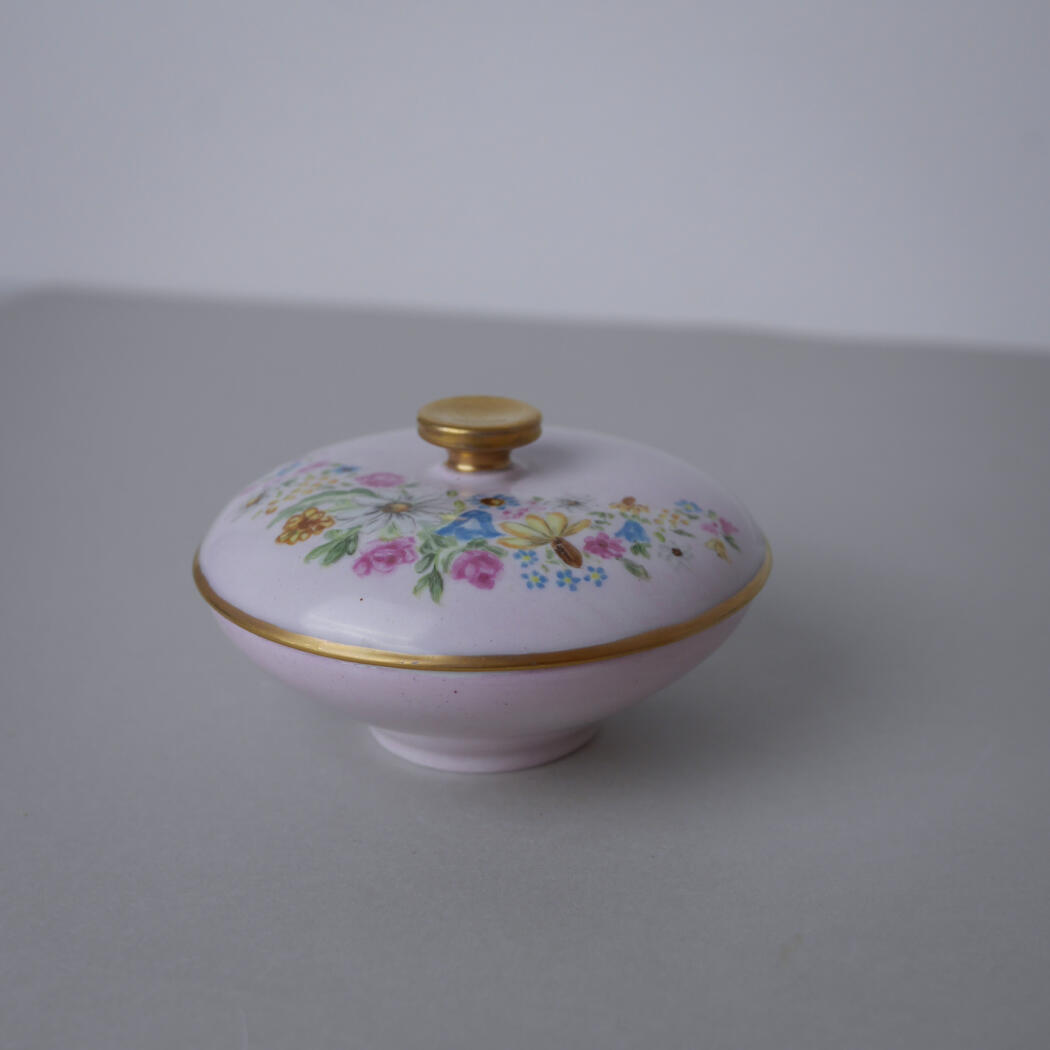 Pink ceramic lidded container with gold trim and a colorful floral pattern on top, resting on a pale surface.