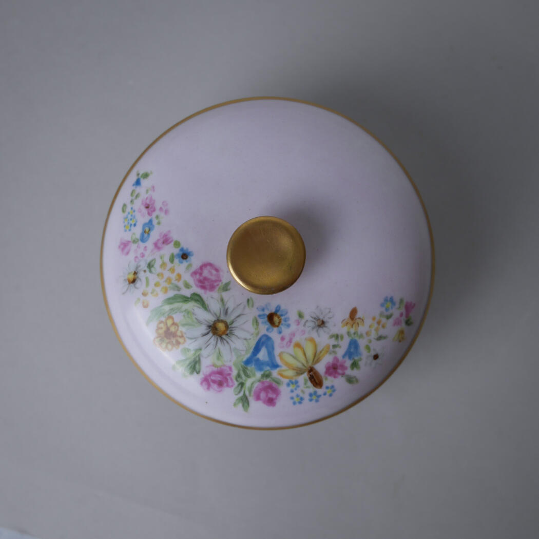 Top-down view of a white porcelain lid with a gold knob and gold rim, adorned with a colorful floral pattern along the edge.