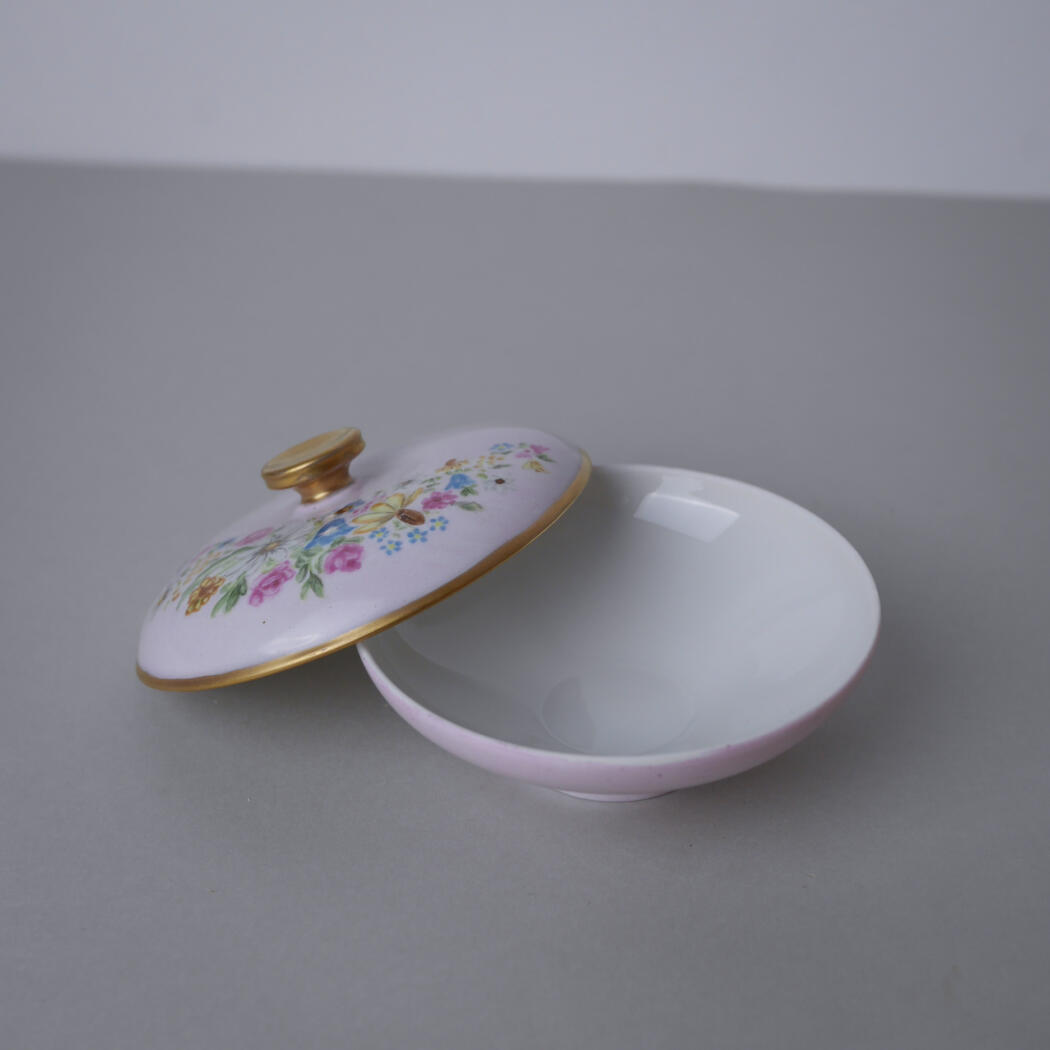 Porcelain round trinket box with a floral lid and gold rim, lid tilted open on a gray surface.