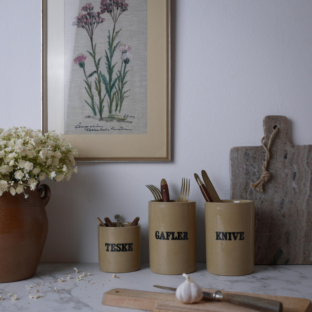 Beige canisters labeled TESKE, GAFLER, KNIVE hold cutlery on a marble counter with a cutting board, garlic, and flowers.