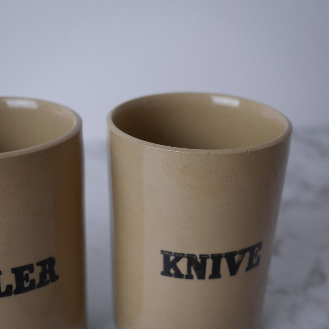 Two beige ceramic mugs with black stamped lettering, partially visible on a light surface.