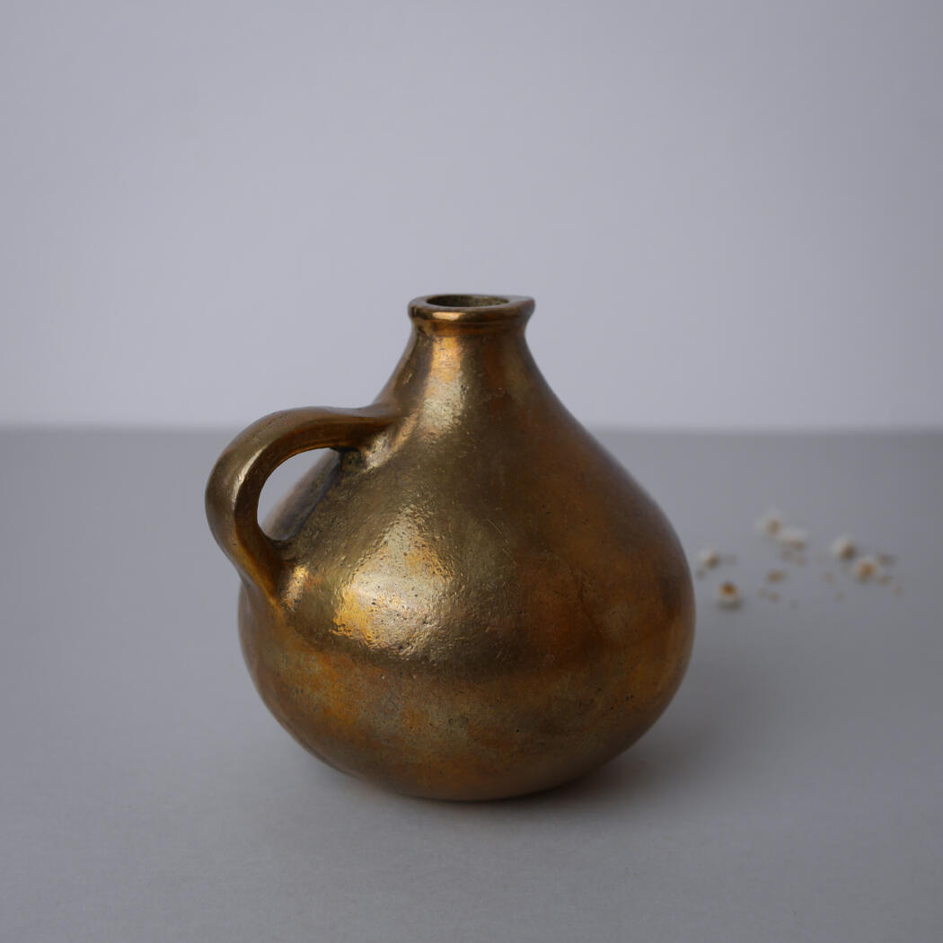 Bronze-colored bulbous ceramic jug with a single handle on a gray surface; small white beads visible to the right side.