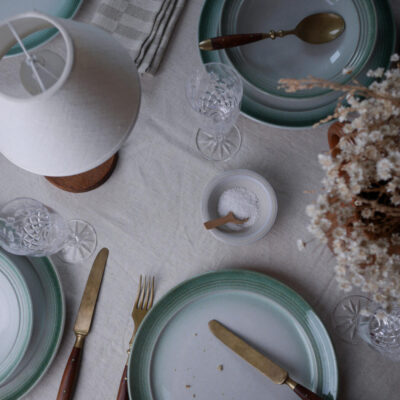 Top-down view of a table setting with mint-green plates, gold flatware, crystal glasses, a salt bowl, and dried flowers on the right.