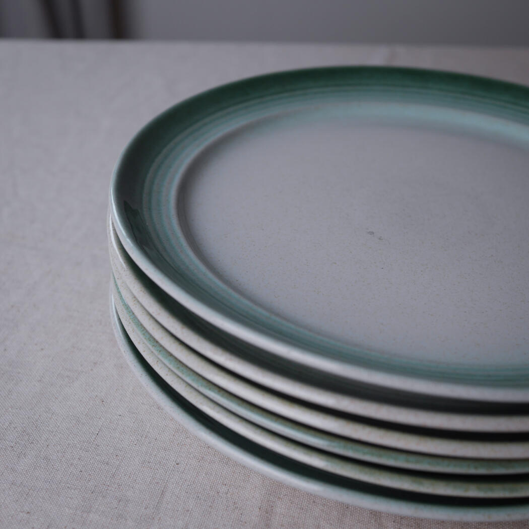 Stack of white plates with a teal-green rim on a light tablecloth, photographed from the side edge.