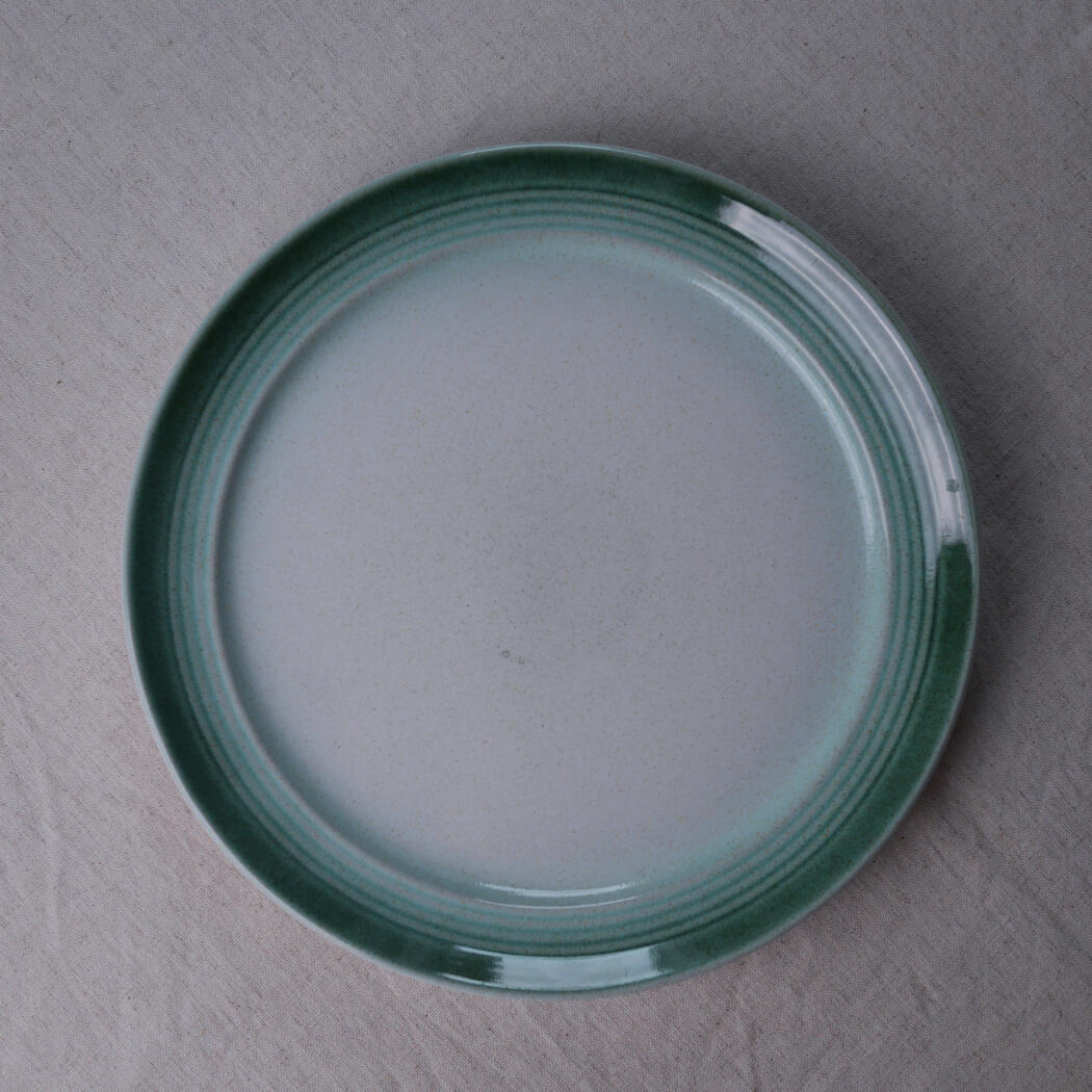 Top-down view of a round ceramic plate with a green glazed rim on a neutral fabric background.