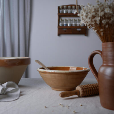 A rustic kitchen scene with two ceramic bowls on a pale wooden table, a wooden spoon resting in the inner bowl, and a rolling pin nearby beside a vase of dried flowers.