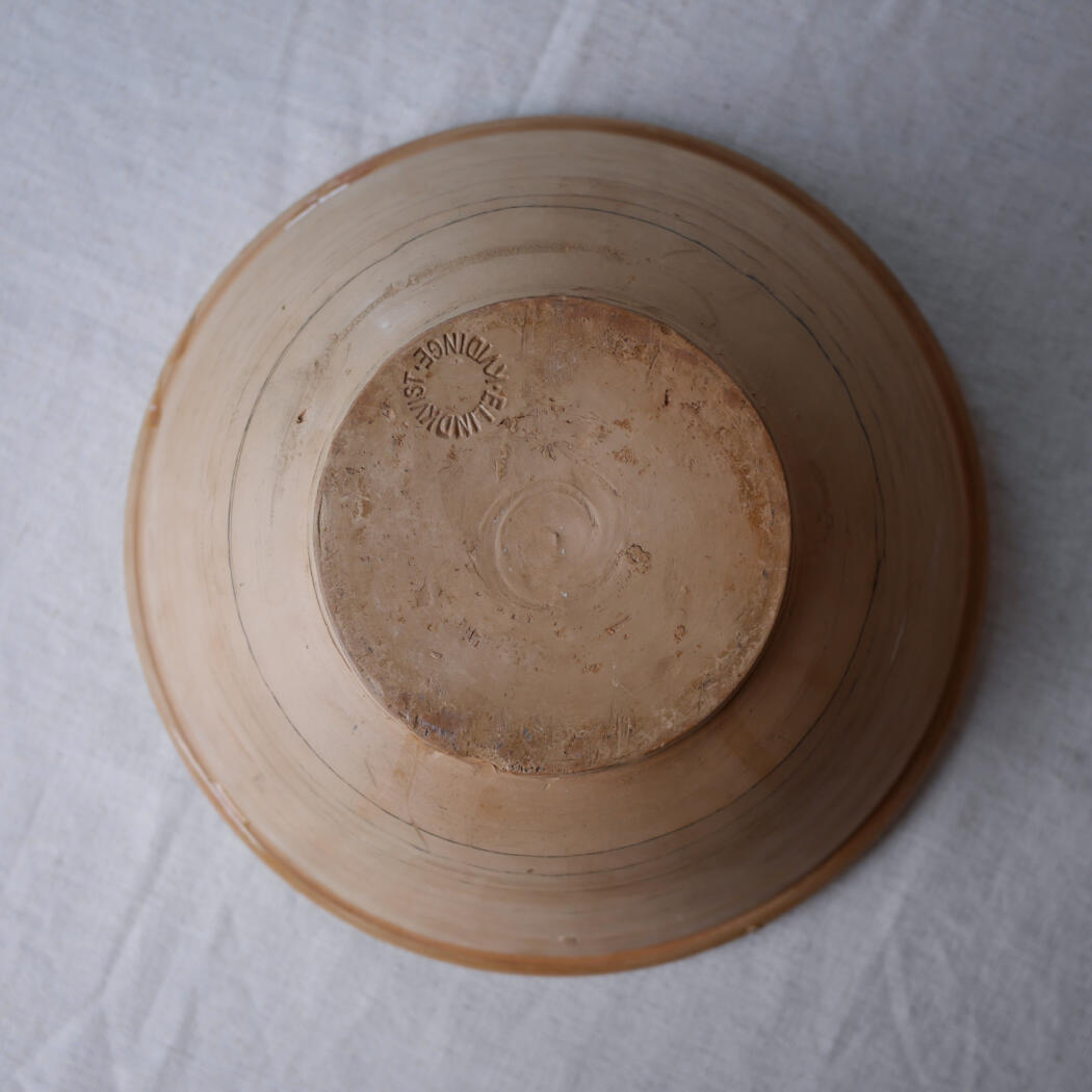 Bottom view of a clay bowl turned upside down on a light fabric surface; circular maker's mark stamped on the base.