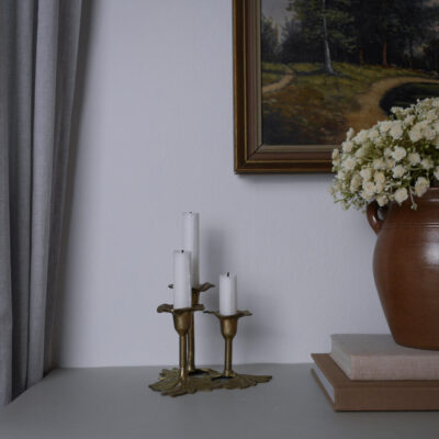 Brass three-candle holder on a gray table with white candles, beside a brown vase of white flowers and stacked books under a landscape painting on the wall.